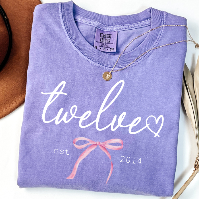 Purple lavender Comfort Colors 12th birthday shirt for girls featuring Twelve script, pink bow, and Est. 2014 design