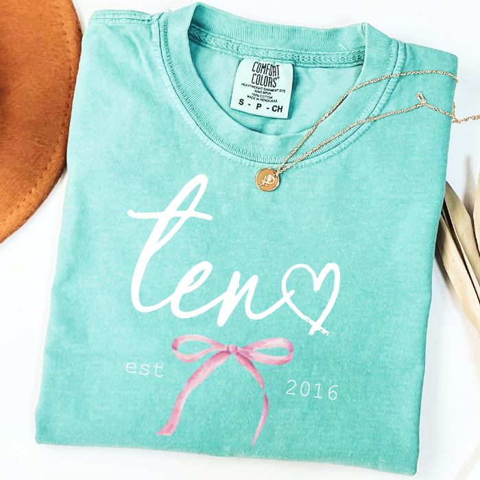 A folded mint green graphic t-shirt with 'ten' in white script, a white heart, and a pink bow coquette, styled with a gold necklace on a white background.