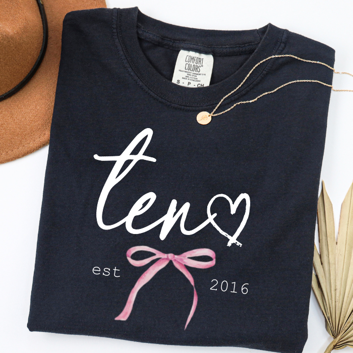 A folded black t-shirt with a white script 'ten' forming a heart, a pink ribbon coquette bow, and 'est 2016' printed below. The shirt is styled with a gold necklace and props like a brown hat and dried palm leaves on a white background.