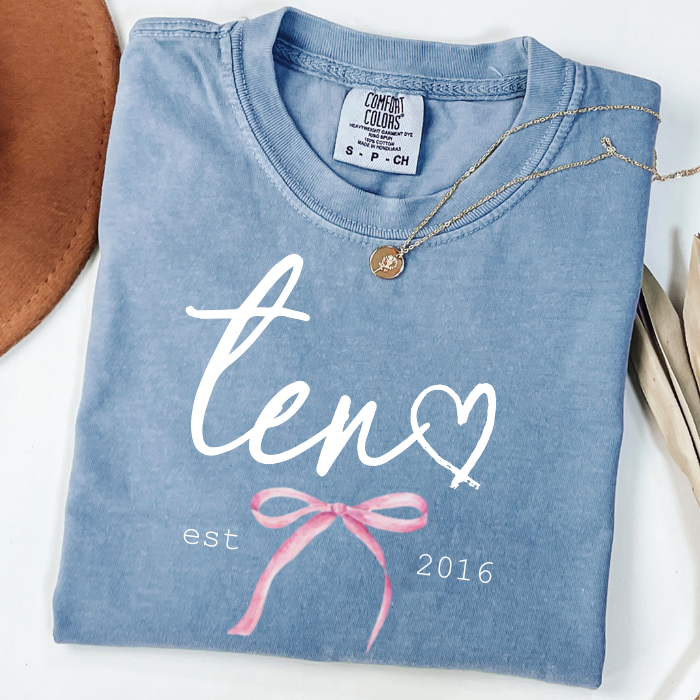A folded light blue t-shirt with a custom white 'ten' script, a heart, and a pink ribbon coquette bow, styled with a gold necklace on a white background.