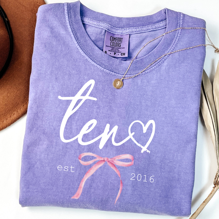 A folded lavender-purple t-shirt with a white script 'ten' and a heart, plus a pink ribbon coquette bow and 'est 2016' text. A gold necklace is draped over the shirt.