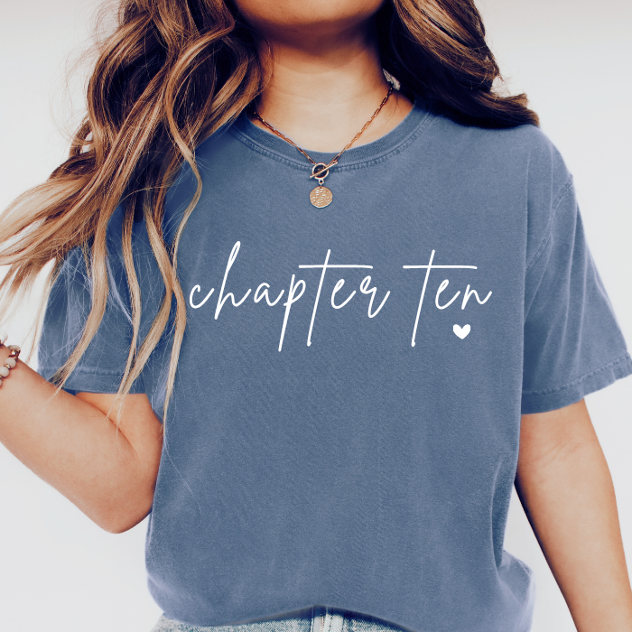 A woman wearing a faded slate blue t-shirt with 'chapter ten' printed in white script across the chest, paired with a gold pendant necklace.