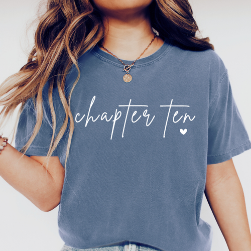 A woman wearing a faded slate blue t-shirt with 'chapter ten' printed in white script across the chest, paired with a gold pendant necklace.