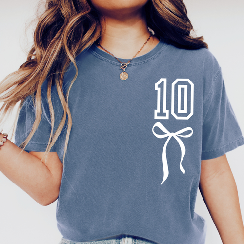 A close-up of a person wearing a slate blue t-shirt with a white graphic print of the number 10 and a bow on the front.