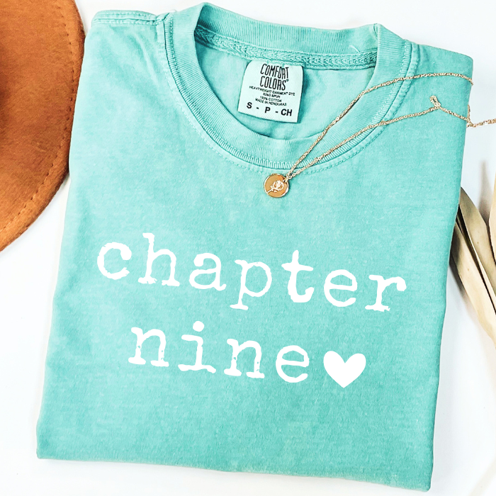 A folded mint green comfort colors t-shirt with 'chapter nine' printed in white text and a white heart, styled with a gold coin necklace on a white background.