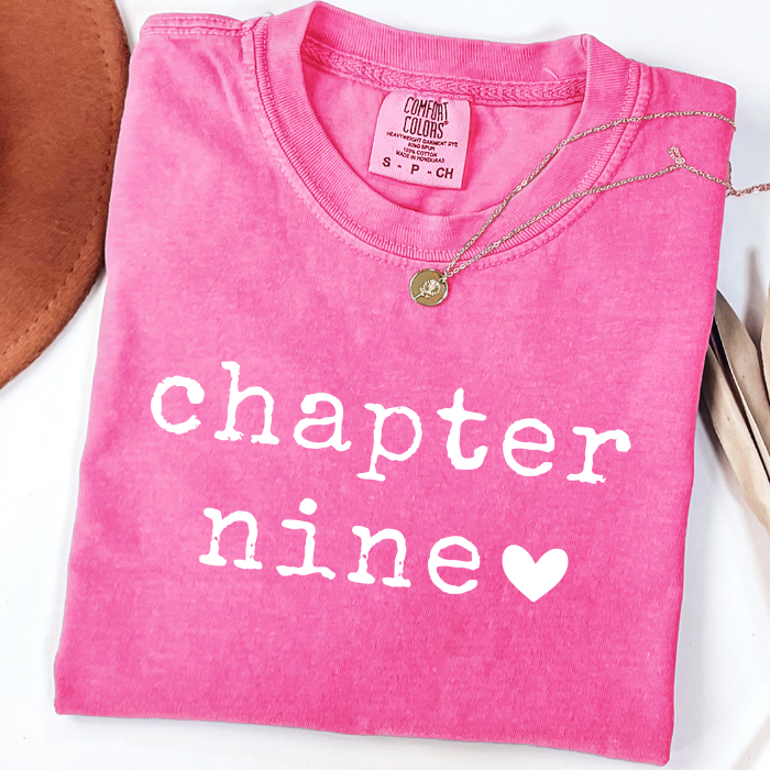A folded bright pink comfort colors t-shirt with 'chapter nine' printed in white text and a white heart, styled with a gold necklace on a white background.