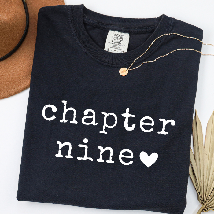 A folded black comfort colors t-shirt with 'chapter nine' printed in white typewriter-style text and a white heart, styled with a gold necklace.