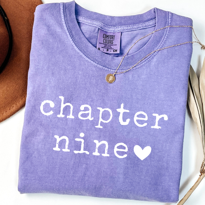 A folded lavender-purple comfort colors t-shirt with 'chapter nine' printed in white text and a white heart, styled with a gold necklace on a white background.