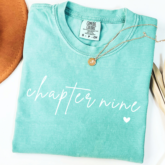 A folded mint green comfort colors t-shirt with 'chapter nine' printed in white script across the front, styled with a gold pendant necklace on a white background.