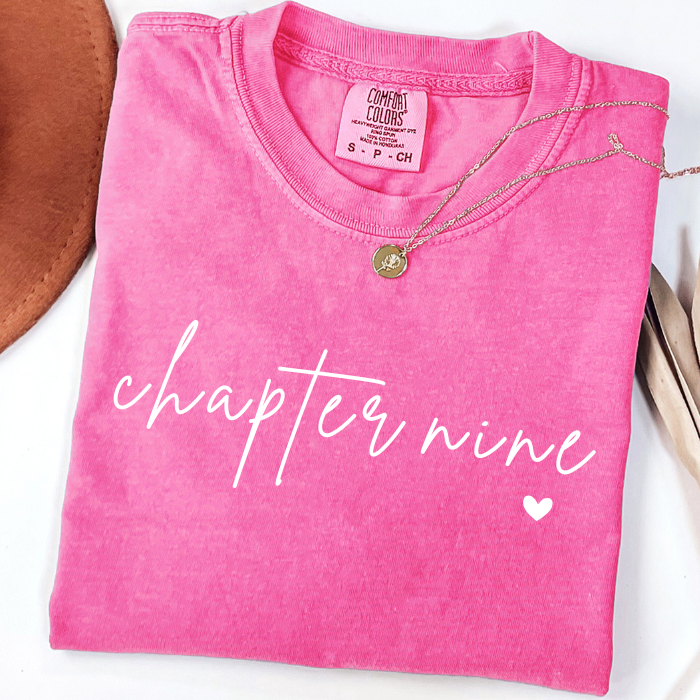 A folded bright pink comfort colors t-shirt with 'chapter nine' printed in white script across the front, styled with a gold necklace on a white background.