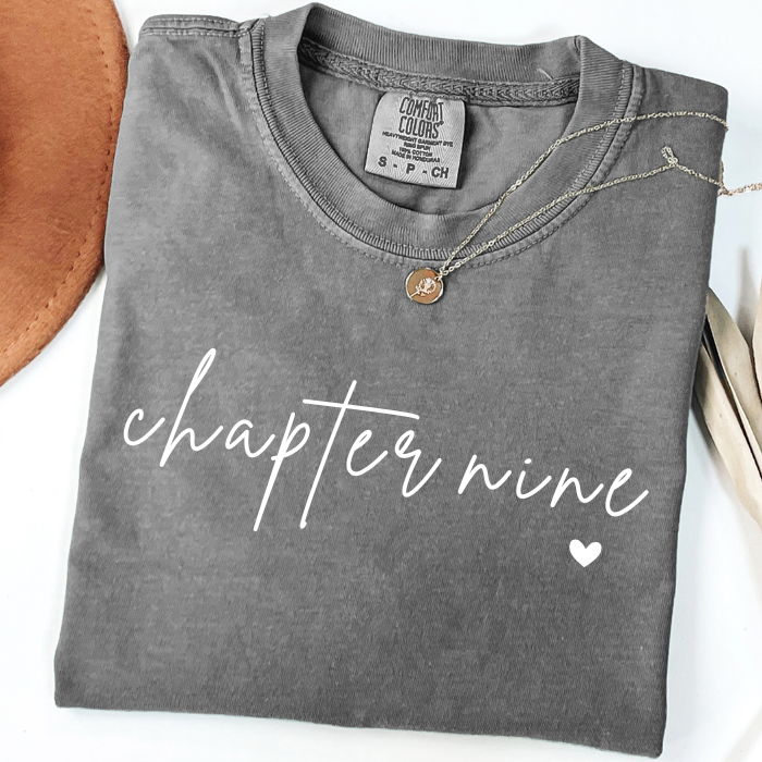 A folded dark gray comfort colors t-shirt with 'chapter nine' printed in white script across the front, styled with a gold necklace on a white background with decorative props.