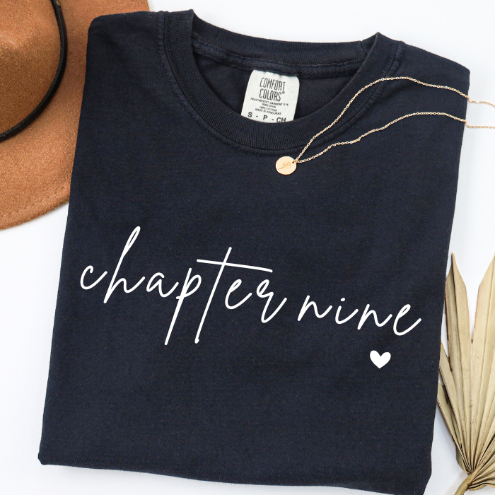 A folded black comfort colors t-shirt with 'chapter nine' written in white script across the front, styled with a gold necklace on a white background with decorative props.