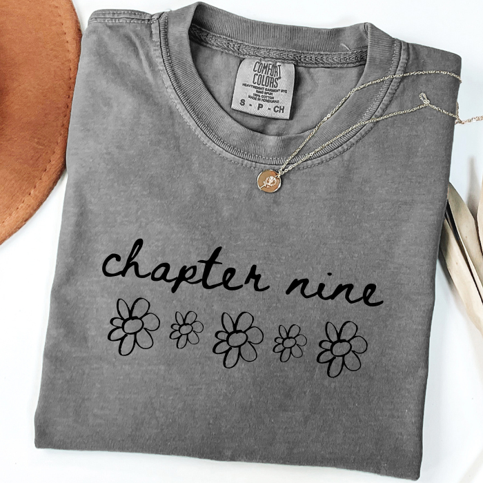 A folded gray comfort colors t-shirt with 'chapter nine' and daisy flower graphics printed on the front, styled with a necklace on a white background with decorative props.