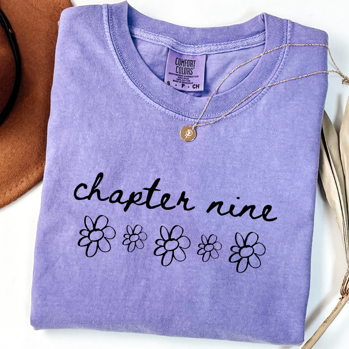 A folded lavender-purple comfort colors t-shirt with 'chapter nine' and daisy flower graphics printed in black on the front. A gold necklace is draped over the collar.