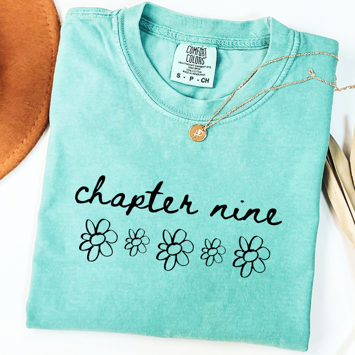 A folded mint green comfort colors t-shirt with 'chapter nine' and daisy flower graphics printed on the front, styled with a gold necklace and a brown leather accessory on a white background.