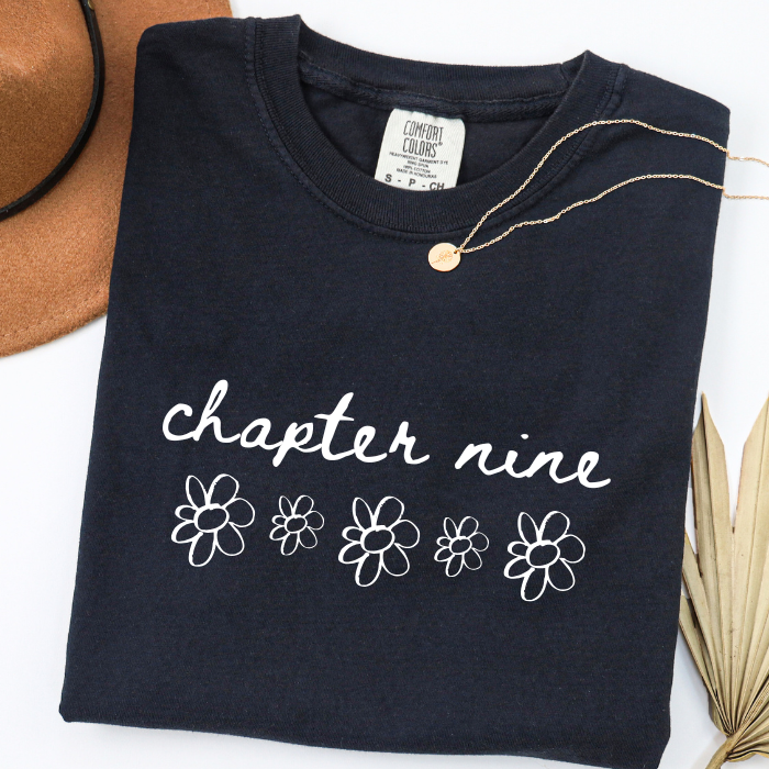 A folded black comfort colors t-shirt with 'chapter nine' and daisy flower graphics in white, styled with a gold necklace, a brown hat, and dried palm leaves on a white background.