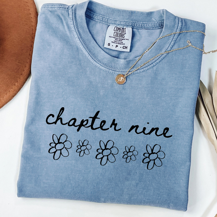A folded light blue comfort colors t-shirt with 'chapter nine' and small daisy flower graphics printed in black on the front. A gold necklace is draped across the collar, and a brown leather-like prop is partially visible on the left, set against a white background.