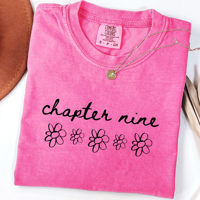 A folded bright pink comfort colors t-shirt with 'chapter nine' and daisy flower graphics printed on the front, styled with a gold necklace on a white background.