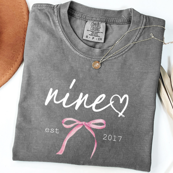 A folded dark gray comfort colors t-shirt with a custom white graphic reading 'nine' and a heart, plus a pink ribbon coquette bow illustration and 'est 2017'. A gold pendant necklace is draped over the collar.