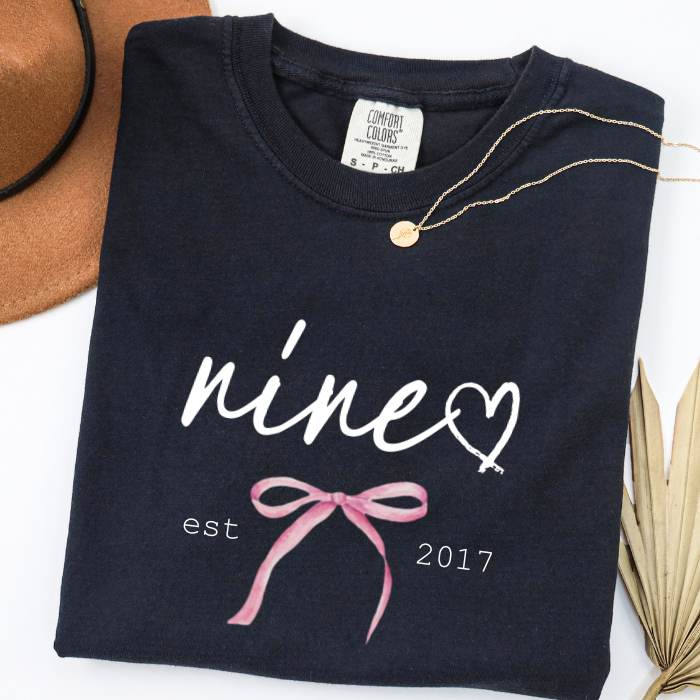 A folded black comfort colors t-shirt with a white 'nine' script, a white heart, a pink coquette bow graphic, and 'est 2017' printed on the front. A gold necklace is draped over the collar, with a brown hat and dried palm leaves partially visible in the background.