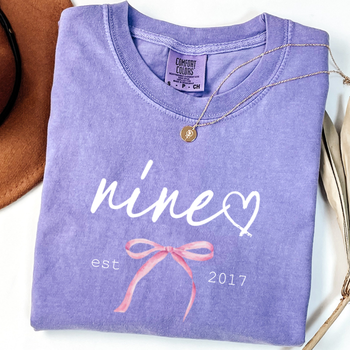 A folded lavender-purple comfort colors t-shirt with a white script graphic reading 'nine' and a pink coquette bow ribbon illustration, styled on a white surface with a gold necklace and a brown hat partially visible.