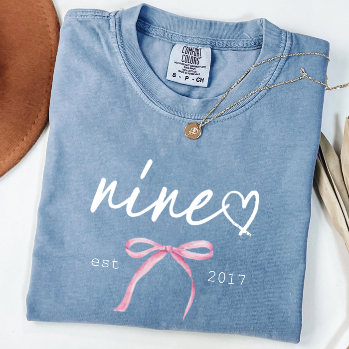 A folded light blue comfort colors t-shirt with a custom white 'nine' and heart graphic, a pink ribbon bow, and 'est 2017' printed on the front. A gold necklace is draped over the collar, and a brown hat is partially visible on the left, all on a white background.