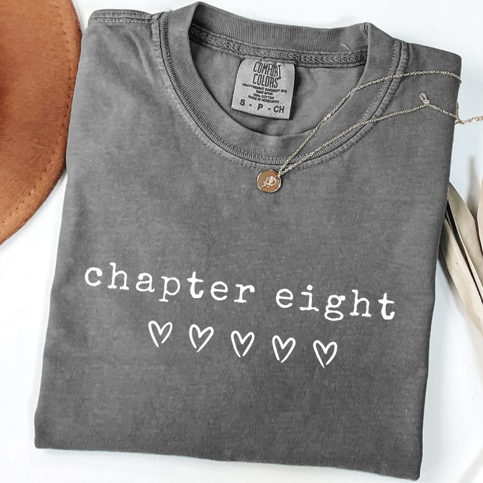 A folded dark gray comfort colors t-shirt with 'chapter nine' printed in white text and a small white heart, styled with a necklace on a white background.