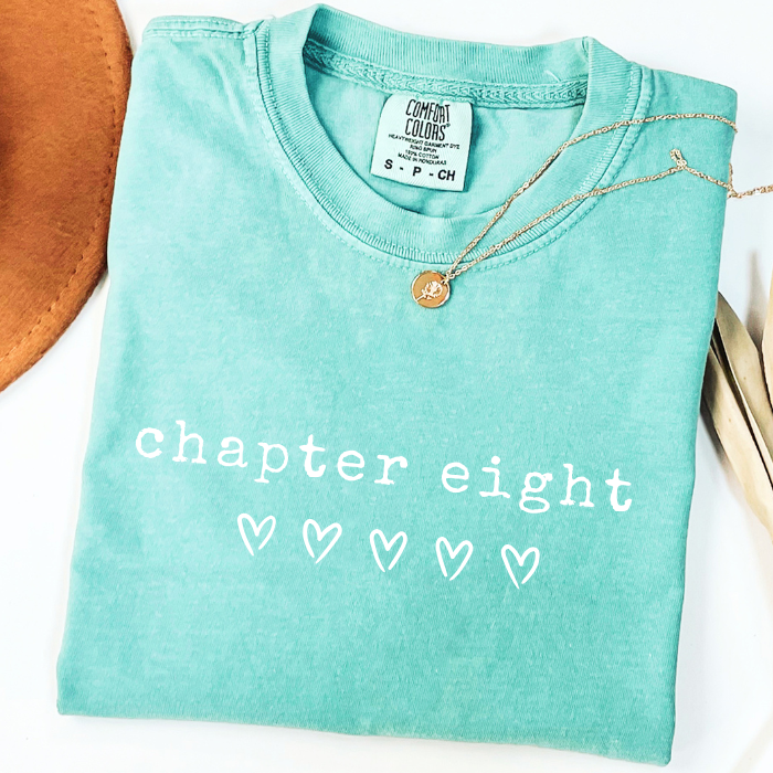 A folded mint green comfort colors t-shirt with 'chapter twelve' printed in white script across the front, styled with a gold pendant necklace and a brown leather accessory on a white background.