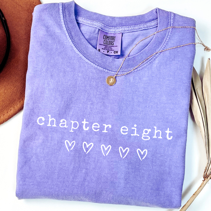 A folded lavender-purple Comfort Colors t-shirt with 'chapter eight' and five heart outlines printed in white on the front. A delicate gold necklace is draped over the collar.