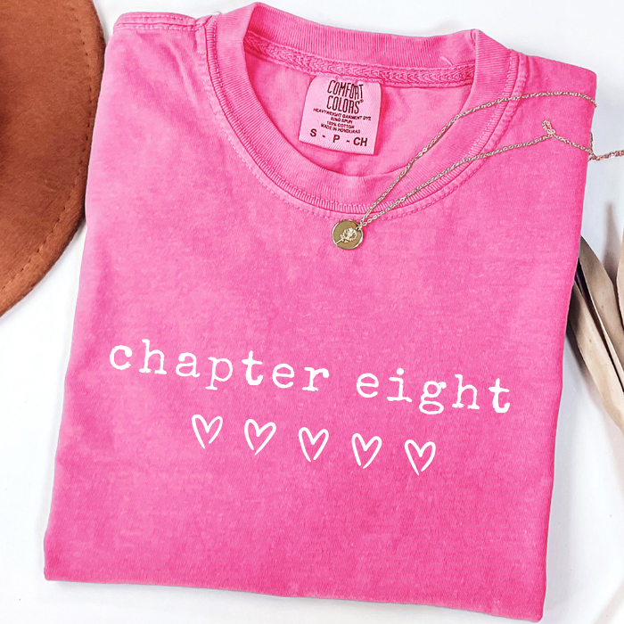 A folded bright pink comfort colors t-shirt with 'chapter eight' and four heart graphics printed in white on the front. A gold necklace is draped over the collar, and a brown leather object is partially visible on the left.