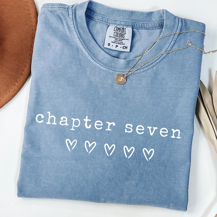 A folded light blue comfort colors t-shirt with 'chapter seven' and five heart outlines printed in white on the front. A delicate gold necklace with a round pendant is draped over the collar.