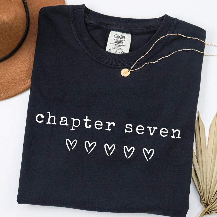 A folded black comfort colors t-shirt with 'chapter seven' and five heart outlines printed in white on the front. A gold necklace is draped over the collar.