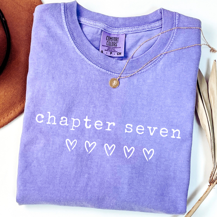 A folded lavender-purple comfort colors Comfort Colors t-shirt with 'chapter seven' and five hearts printed in white on the front, styled with a gold necklace and a brown hat on a white background.