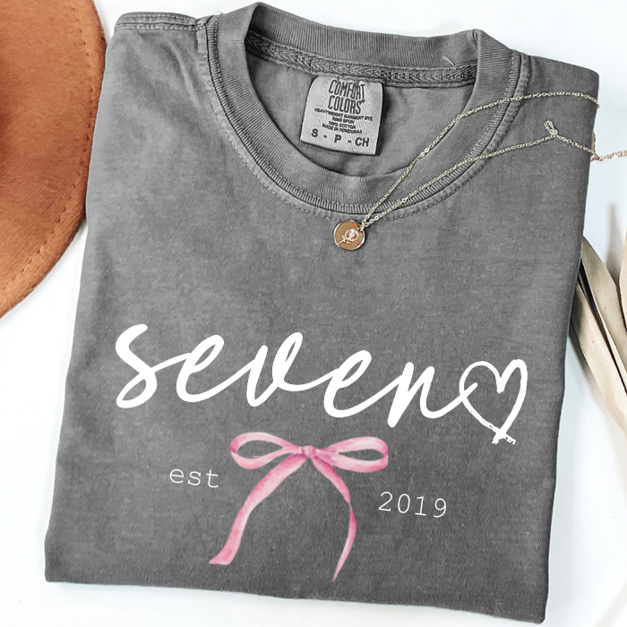 A folded dark gray comfort colors t-shirt with a graphic print that reads 'seven est 2019' in white, decorated with a pink bow illustration and a white heart. A gold pendant necklace is draped over the collar.