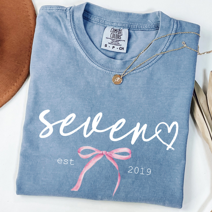 A folded light blue comfort colors t-shirt with 'seven' and 'est 2019' printed in white, decorated with a pink bow graphic. A gold necklace is draped over the collar, and the shirt is styled with a brown hat on a white background.