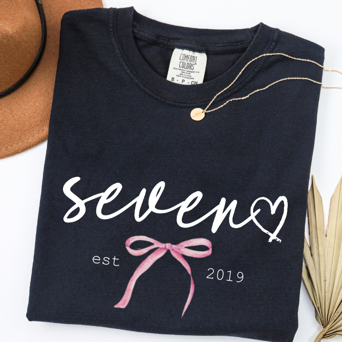 A folded black comfort colors t-shirt with a graphic reading 'seven est 2019' and a pink ribbon, styled on a white background with a gold necklace, a brown hat, and dried palm leaves.