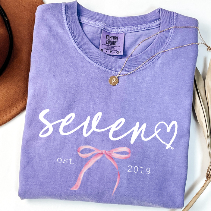 A folded lavender Comfort Colors t-shirt with a custom graphic print reading 'seven est 2019' in white script and a pink ribbon illustration. A gold necklace is draped over the collar, and a brown hat and dried leaves are partially visible as styling props on a white surface.