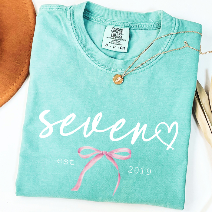 A folded mint green comfort colors t-shirt with 'seven' and 'est 2019' printed in white, decorated with a pink bow graphic. A gold necklace is draped over the collar, and a brown leather hat is partially visible on the left, all on a white background.