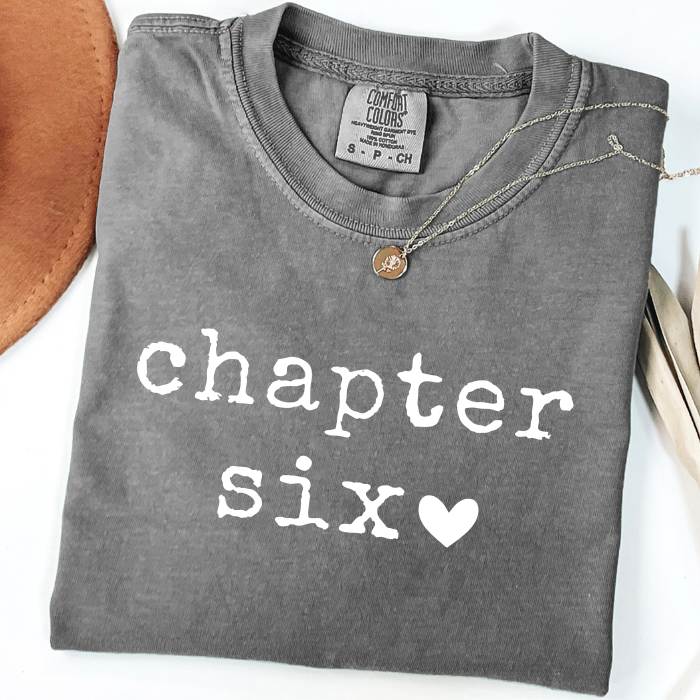A folded dark gray comfort colors t-shirt with 'chapter six' printed in white text and a small white heart, styled with a necklace on a white background.