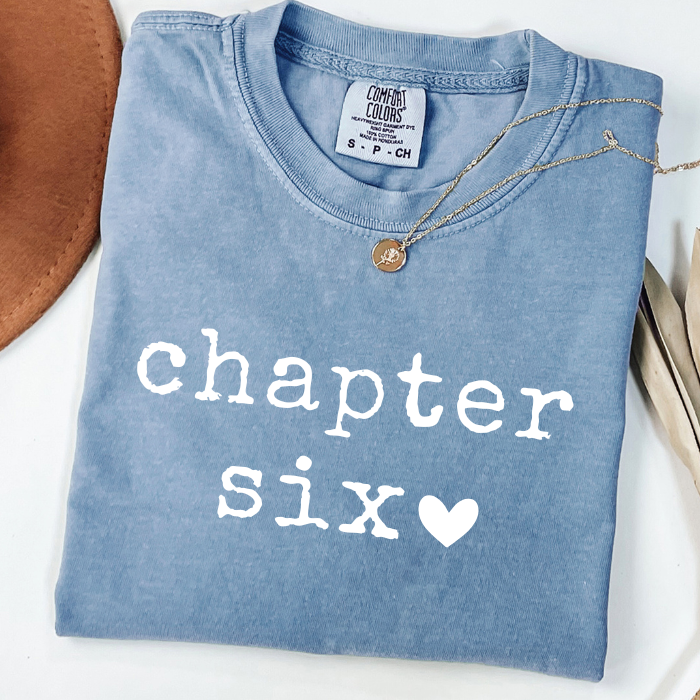A folded light blue comfort colors t-shirt with 'chapter six' printed in white typewriter-style text and a white heart, styled with a delicate gold necklace on a white background.