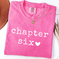 A folded bright pink comfort colors t-shirt with 'chapter six' printed in white text and a white heart, styled with a gold necklace on a white background.