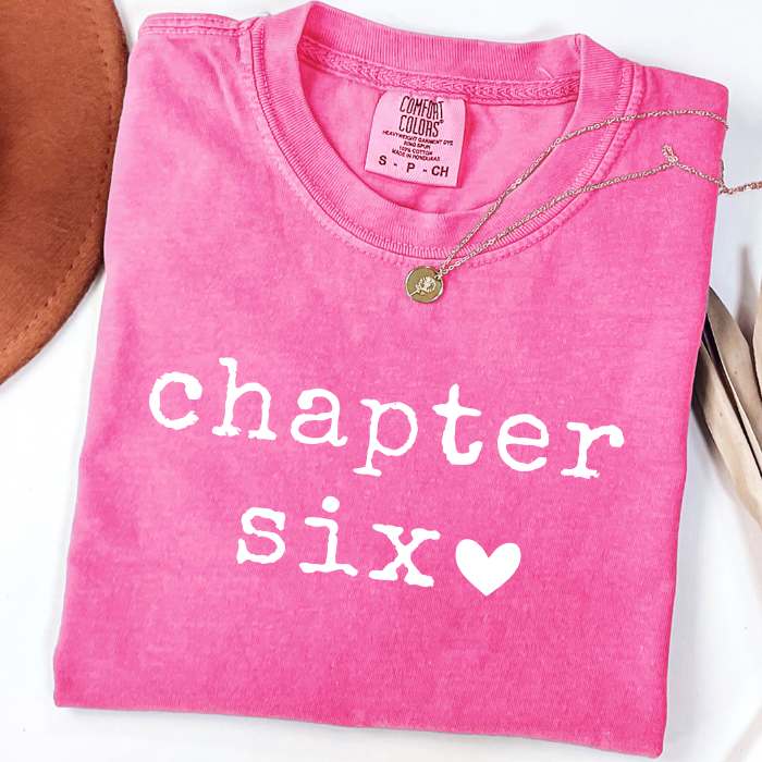 A folded bright pink comfort colors t-shirt with 'chapter six' printed in white text and a white heart, styled with a gold necklace on a white background.