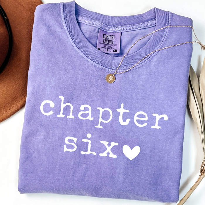 A folded lavender-purple comfort colors t-shirt with 'chapter six' printed in white text and a white heart, styled with a gold necklace on a white background.