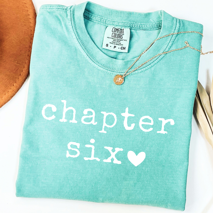 A folded mint green comfort colors t-shirt with 'chapter six' printed in white text and a small white heart, styled with a gold necklace on a white background.