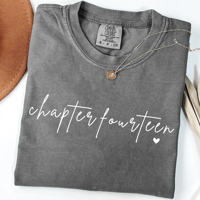 A folded dark gray comfort colors t-shirt with 'chapter fourteen' printed in white script across the front, styled with a small bronze pendant necklace on a white background with decorative props.