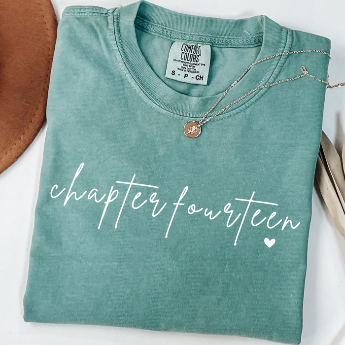 A folded sage green comfort colors t-shirt with 'chapter fourteen' printed in white script across the front, styled with a silver necklace and a brown leather accessory on a white background.