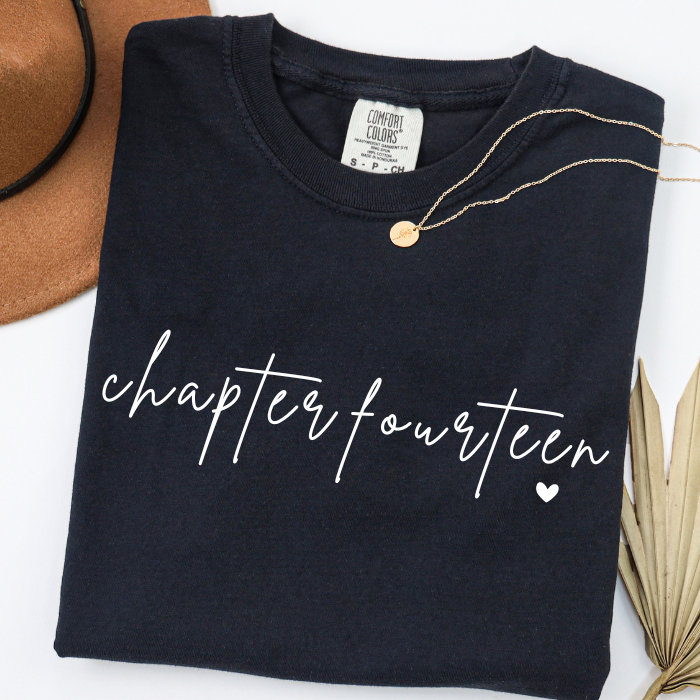 A folded black comfort colors t-shirt with 'chapter fourteen' written in white script across the front, styled with a gold necklace, a brown hat, and dried palm leaves on a white background.