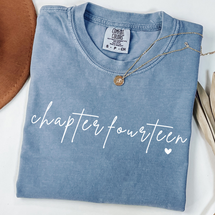 A folded light blue comfort colors t-shirt with 'chapter fourteen' printed in white script across the front, styled with a gold necklace and props on a white background.