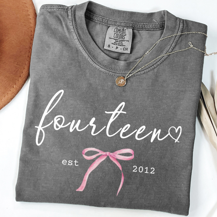 A folded dark gray comfort colors t-shirt with 'fourteen est 2012' printed in white and pink, styled with a gold pendant necklace on a white background.