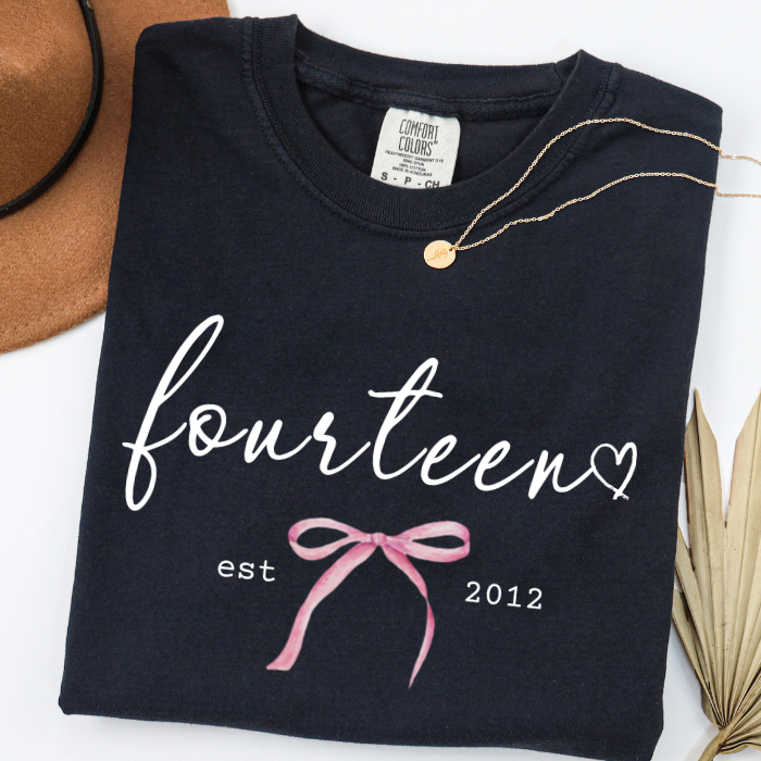 A folded black comfort colors t-shirt with 'fourteen' in white script, 'est 2012' in white, and a pink bow graphic. A gold necklace is draped over the collar, with a brown hat and dried palm leaves partially visible in the background.